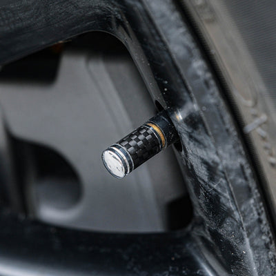 Close-up of a carbon fiber car tire valve stem cap with a checkered pattern and metallic detailing, installed on a vehicle's wheel rim
