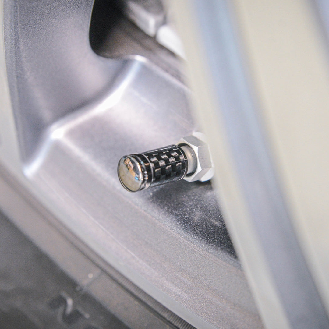 Close-up of a carbon fiber car tire valve stem cap with a checkered pattern, installed on a silver vehicle wheel rim.