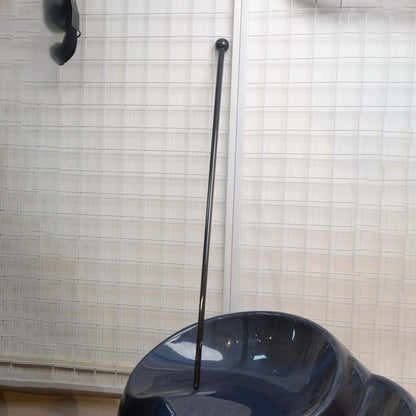 Close-up of a full-length carbon fiber cane with a spherical top, resting on a carbon fiber object against a grid-patterned wall.