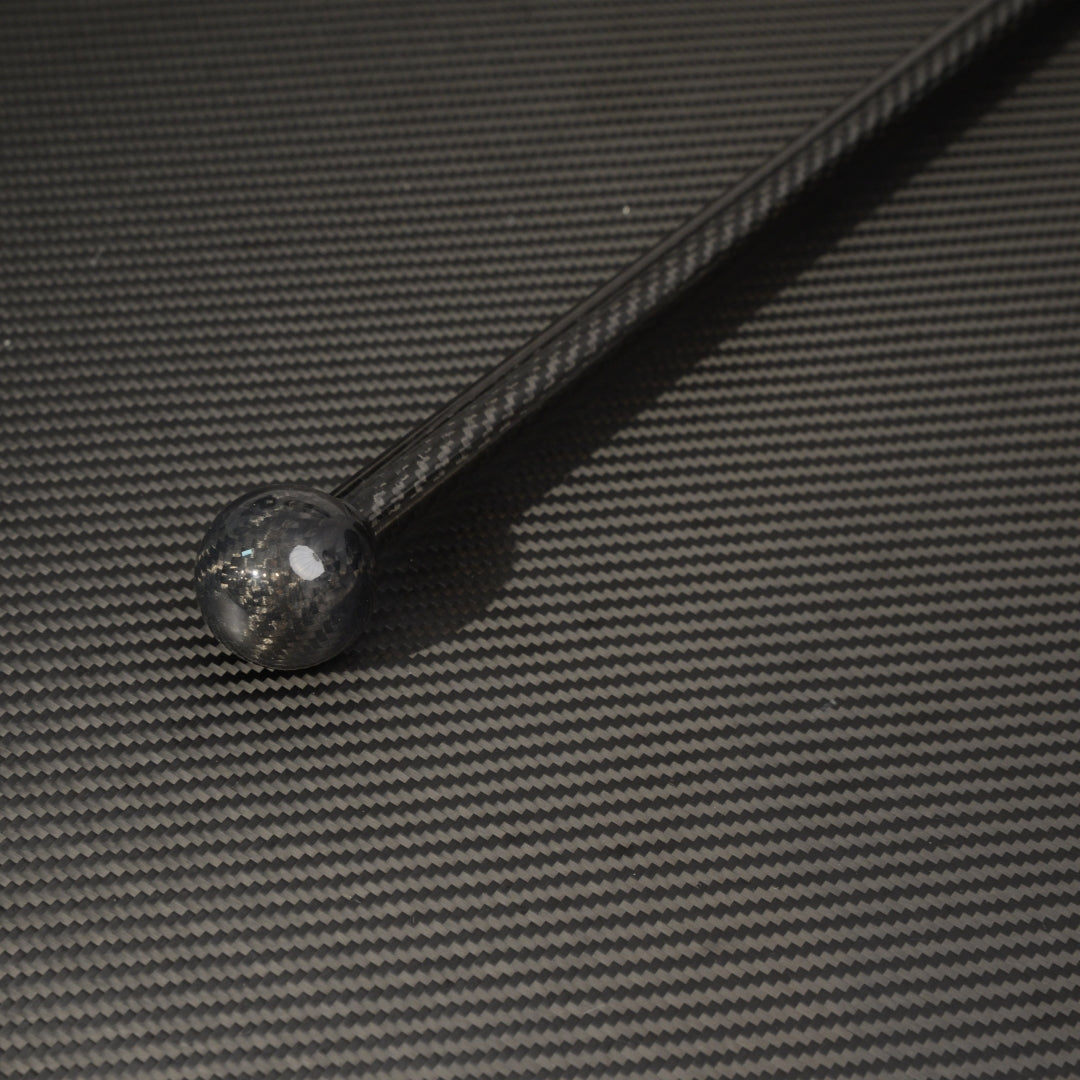 Close-up of a full-length carbon fiber cane with a spherical top, showcasing the carbon fiber weave pattern, placed on a carbon fiber textured surface.