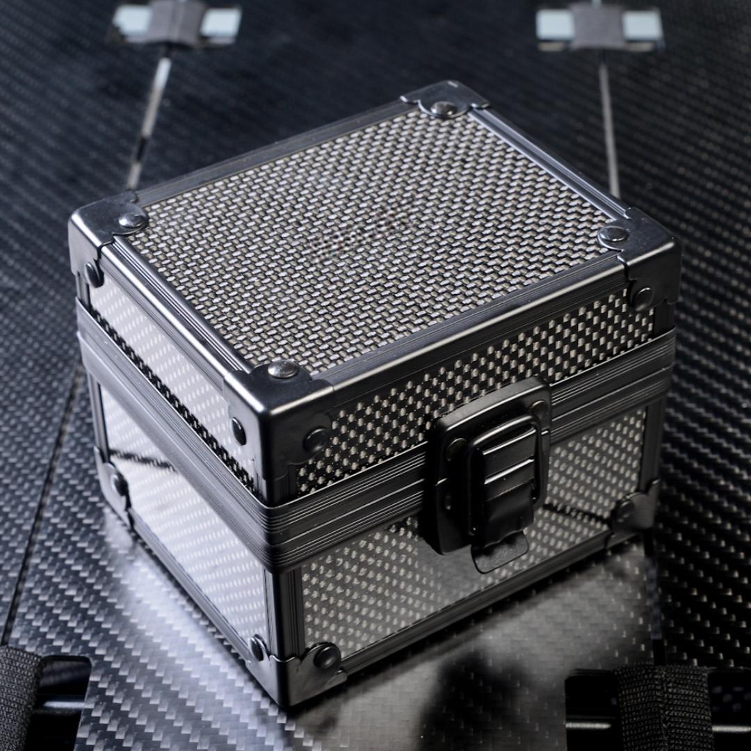 Close-up of a square 3K carbon fiber watch box with a checkered pattern, metallic corner reinforcements, and a black latch, placed on a carbon fiber textured surface.