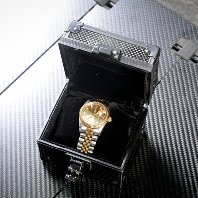 Close-up of an open 3K carbon fiber watch box with a checkered pattern, featuring a black velvet interior and a gold wristwatch inside