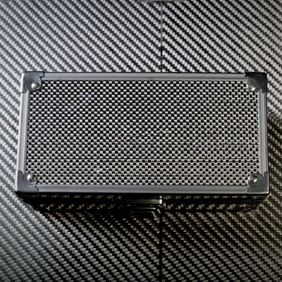 Close-up of a rectangular 3K carbon fiber watch box with a checkered pattern and metallic corner accents, placed on a carbon fiber textured surface.