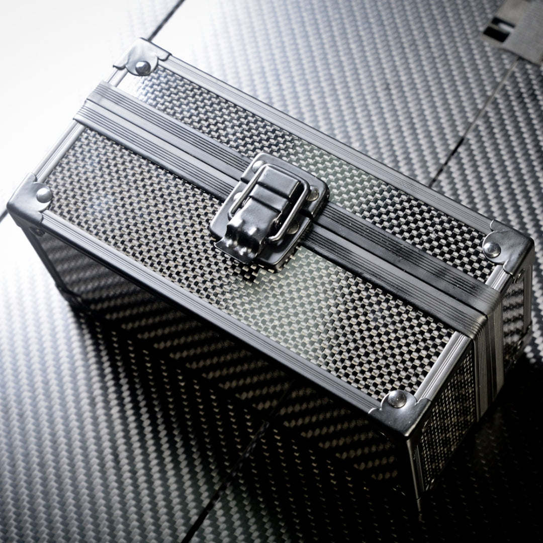 Close-up of a rectangular 3K carbon fiber watch box featuring a checkered pattern, a metallic latch, and metallic corner accents, positioned on a carbon fiber textured surface.
