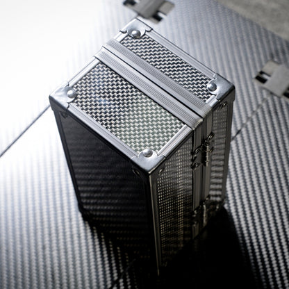 Close-up of a cube-shaped 3K carbon fiber watch box with a checkered pattern and metallic corner reinforcements, positioned on a carbon fiber textured surface.