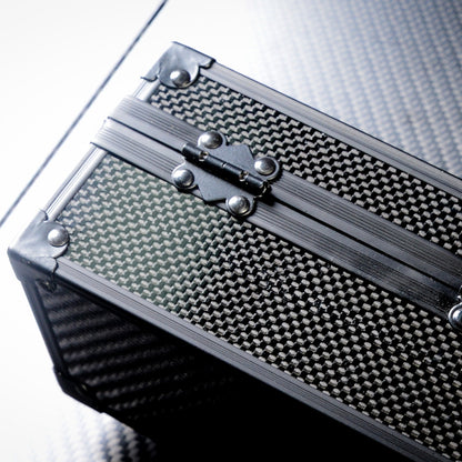 Close-up of a 3K carbon fiber watch box featuring a checkered pattern, a metallic hinge with rivets, and reinforced corners, positioned on a sleek surface.