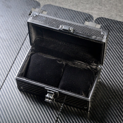 Close-up of an open 3K carbon fiber watch box with a black velvet interior featuring two cushioned slots for watches, metallic latches