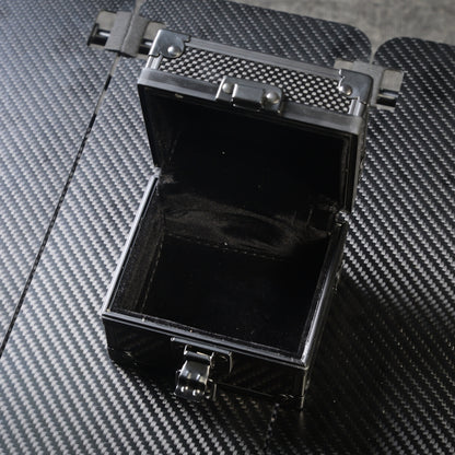 Close-up of an open 3K carbon fiber watch box with a black velvet interior, metallic latches, and a carbon fiber textured exterior, placed on a matching carbon fiber surface.