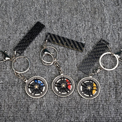 Close-up of three carbon fiber keychains featuring wheel rim designs with colorful brake calipers (blue, red, and yellow), along with carbon fiber-textured accents (including a strip with a ruler scale), displayed on a gray textured fabric surface.