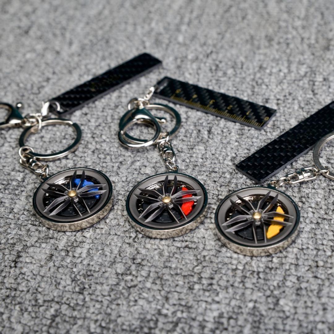 Close-up of three carbon fiber keychains featuring wheel rim designs with colorful brake calipers (blue, red, and yellow), accompanied by carbon fiber-textured accents (including a ruler-marked piece), displayed on a gray textured fabric surface.