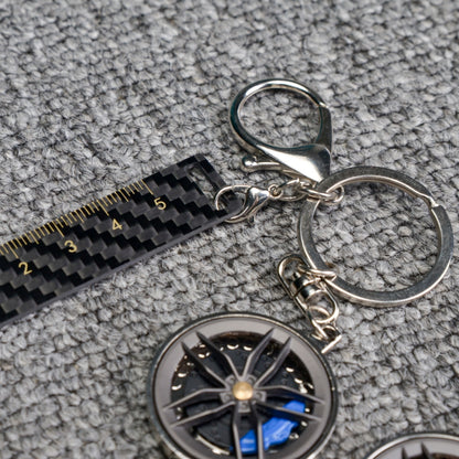 Close-up of a carbon fiber keychain featuring a wheel rim design with blue brake calipers, along with a carbon fiber-textured ruler accent, displayed on a gray textured fabric surface.