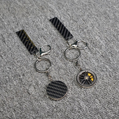 Close-up of two carbon fiber keychains—one featuring a wheel rim design with yellow brake calipers, and the other with a carbon fiber circular accent—both accompanied by carbon fiber-textured strips (one including a ruler scale), displayed on a gray textured fabric surface.