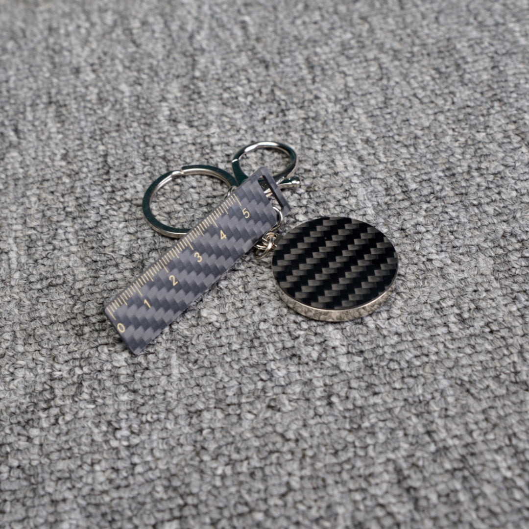 Close-up of a carbon fiber keychain featuring a ruler - shaped accent with measurement markings and a circular carbon fiber charm, displayed on a gray textured fabric surface.