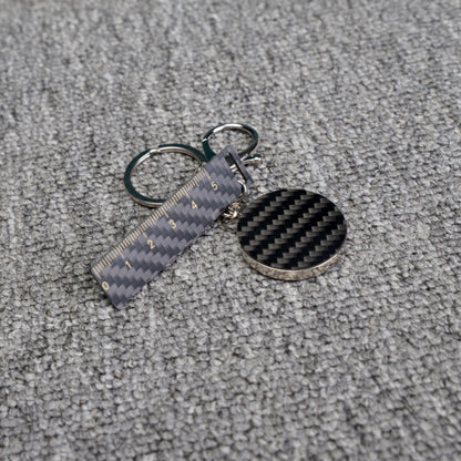 Close-up of a carbon fiber keychain featuring a ruler - shaped accent with measurement markings and a circular carbon fiber charm, displayed on a gray textured fabric surface.