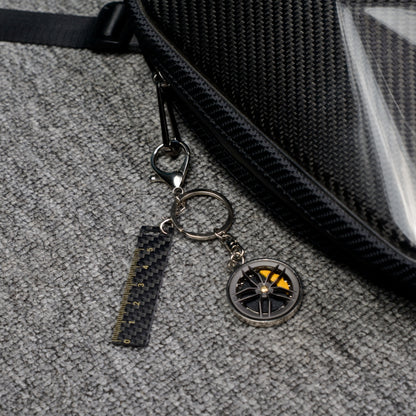Close-up of a carbon fiber keychain featuring a wheel rim charm with yellow brake calipers, a carbon fiber ruler - shaped accent with measurement markings, attached to a carbon fiber - textured bag, displayed on a gray textured fabric surface.