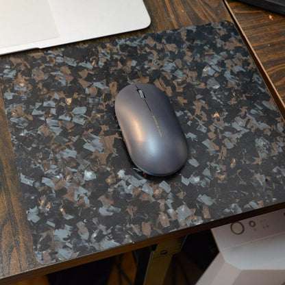 "A sleek gray wireless mouse resting on a forged carbon fiber mouse pad with a textured, fragmented black and brown pattern, situated on a wooden desk with a laptop partially visible in the background."