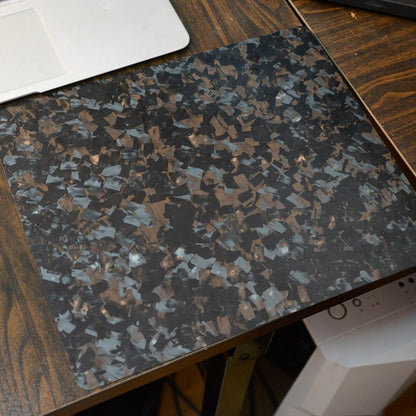 "A forged carbon fiber mouse pad with a textured, fragmented black and brown pattern, placed on a wooden desk with a partial view of a laptop in the background."