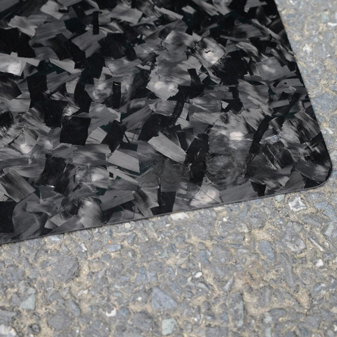 Close-up of a forged carbon fiber mouse pad with a textured, fragmented black and gray pattern, positioned on a rough asphalt surface.
