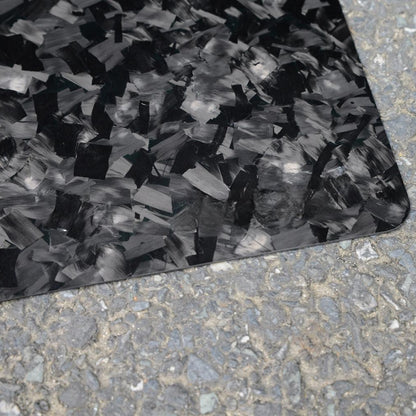 Close-up of a forged carbon fiber mouse pad with a textured, fragmented black and gray pattern, positioned on a rough asphalt surface.