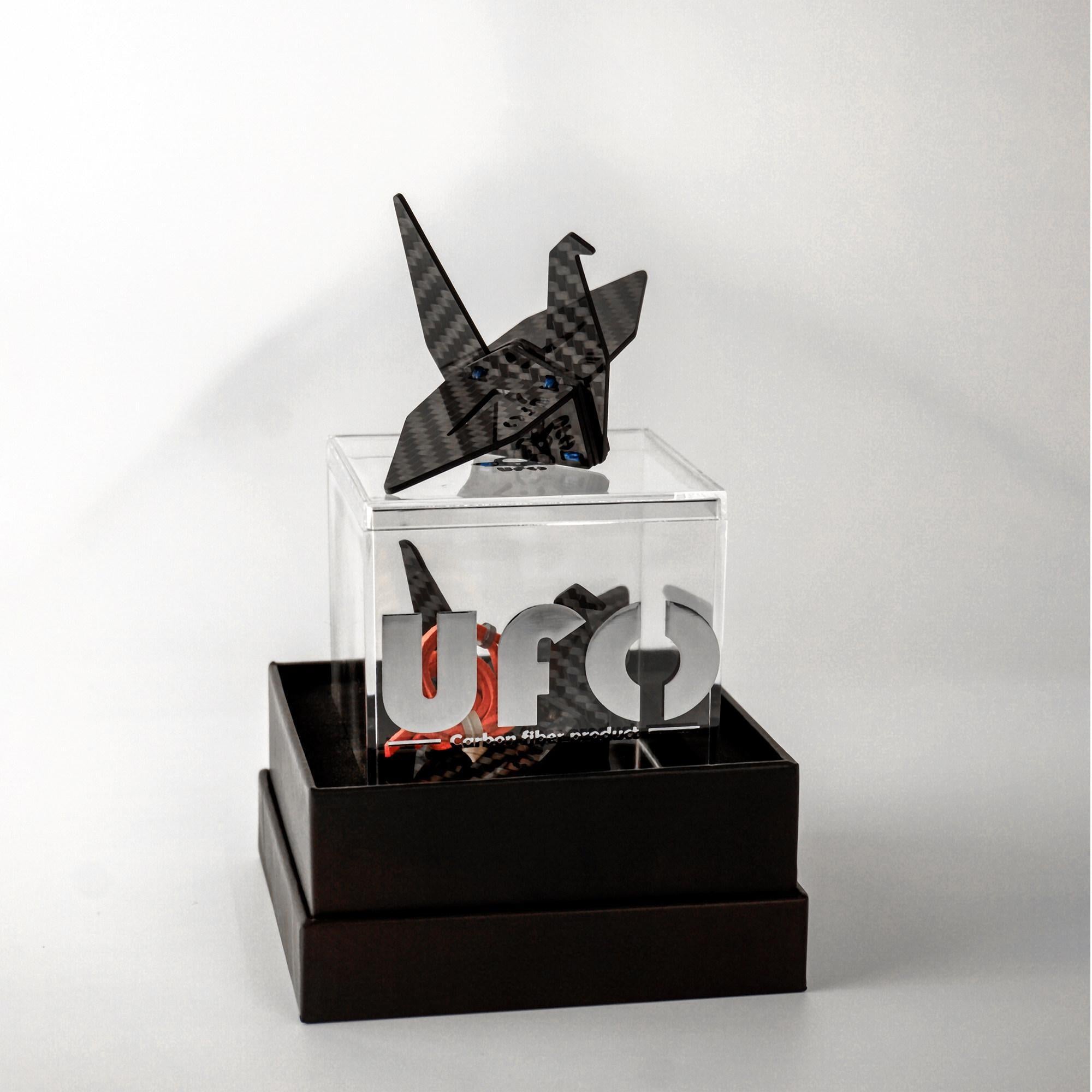 "Black carbon fiber-textured origami crane (with small blue accent details) placed in a clear acrylic case (featuring 'UFO' branding and 'Carbon Fiber product' text); the acrylic case sits on a dark double-layered box, set against a light white background"