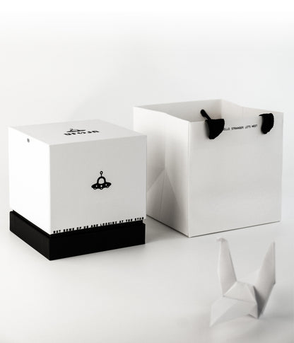 "Minimalist black-and-white packaging set: a square gift box (with a black base) and a matching paper bag. Both feature a small black UFO illustration — the box has the phrase 'BUT SOME OF US ARE LOOKING AT THE STAR' along its bottom edge, while the bag displays 'HELLO, STRANGER. LET'S MEET' near its black fabric handles. A white origami crane sits beside the packaging, emphasizing the clean, modern aesthetic."