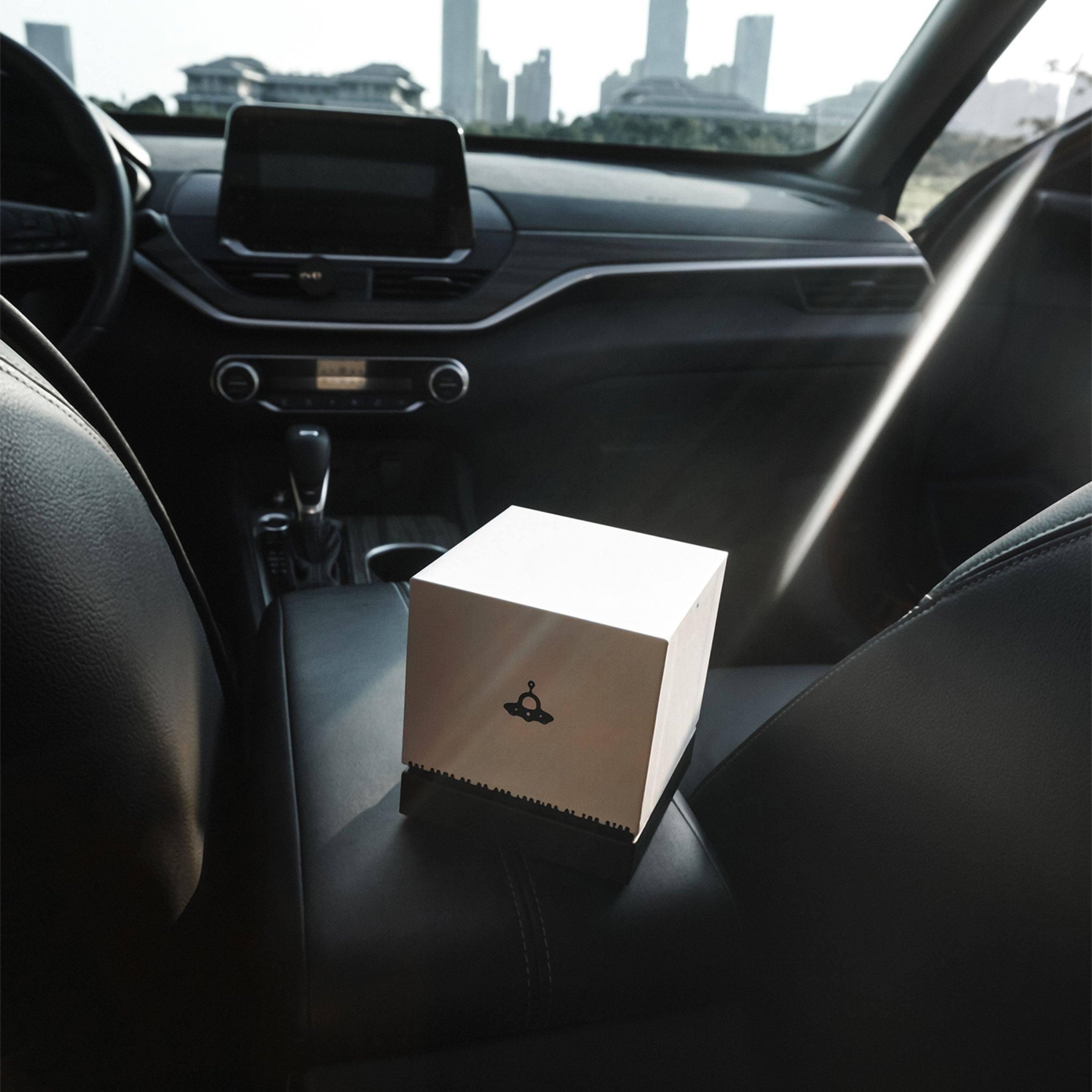 "Interior of a modern car with black leather seating: A small white cube-shaped box (decorated with a black UFO graphic and perched on a black base) sits on the center console area. The car’s dashboard, infotainment screen, and gear shift are visible in the background; bright daylight (with a noticeable light beam) and city skyscrapers can be seen through the vehicle’s windows."
