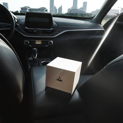 "Interior of a modern car with black leather seating: A small white cube-shaped box (decorated with a black UFO graphic and perched on a black base) sits on the center console area. The car’s dashboard, infotainment screen, and gear shift are visible in the background; bright daylight (with a noticeable light beam) and city skyscrapers can be seen through the vehicle’s windows."
