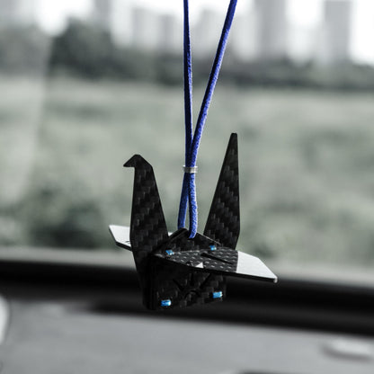 "Black carbon fiber-textured origami crane car charm (featuring small blue accent rivets), suspended from a vibrant blue cord; displayed inside a vehicle with a softly blurred outdoor landscape in the background (minimalist modern decorative accessory)"