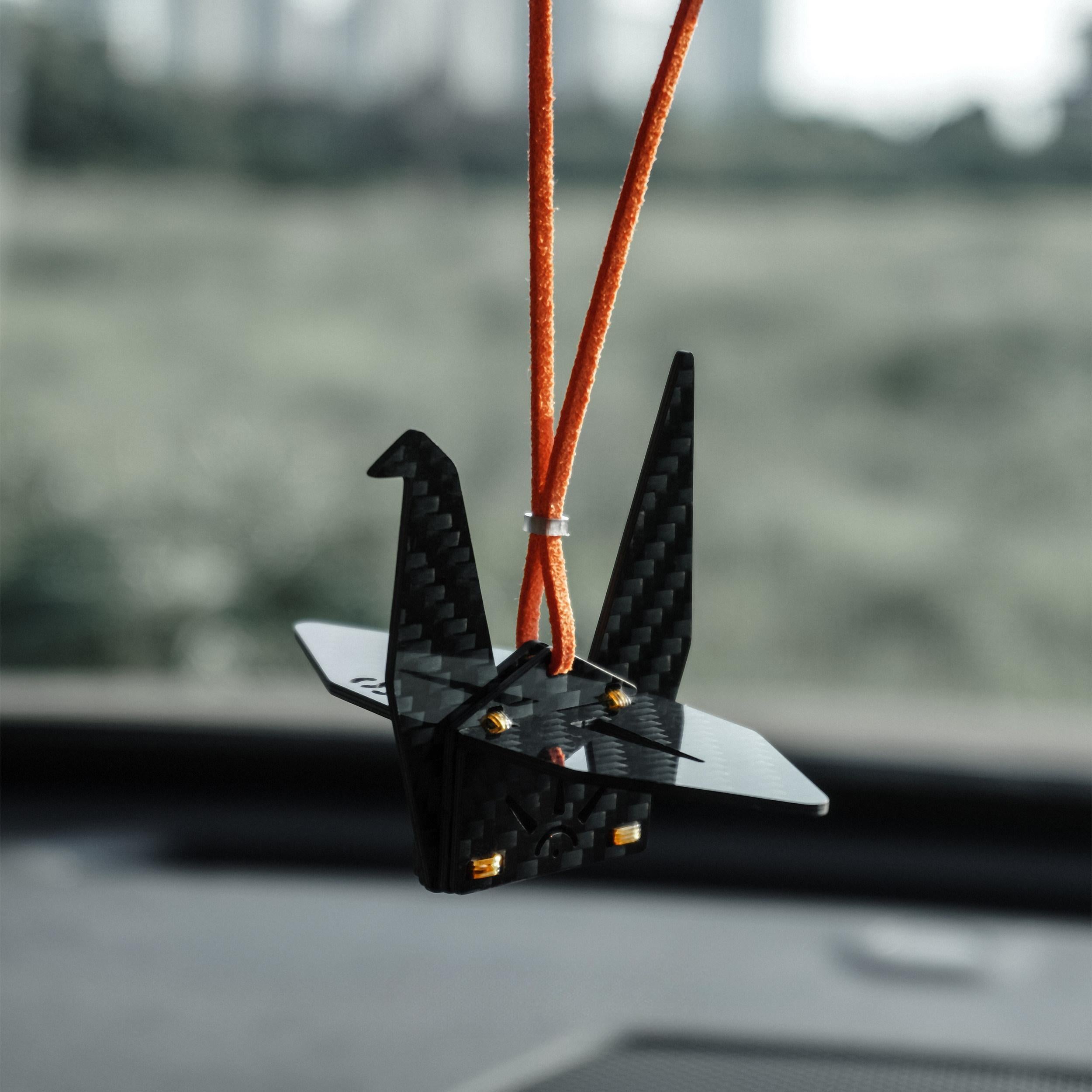"Black carbon fiber-textured origami crane car charm (adorned with small gold accent rivets), suspended from a bright orange cord; displayed inside a vehicle against a softly blurred outdoor landscape (minimalist modern decorative accessory)"
