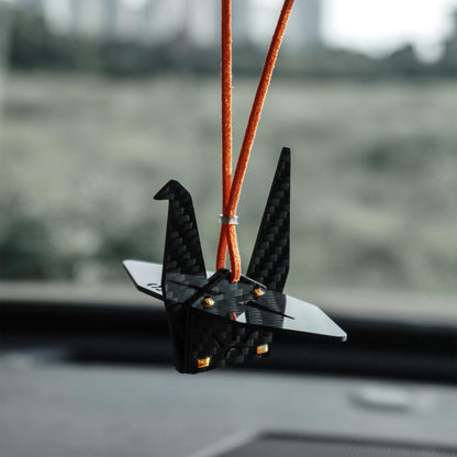 "Black carbon fiber-textured origami crane car charm (adorned with small gold accent rivets), suspended from a bright orange cord; displayed inside a vehicle against a softly blurred outdoor landscape (minimalist modern decorative accessory)"