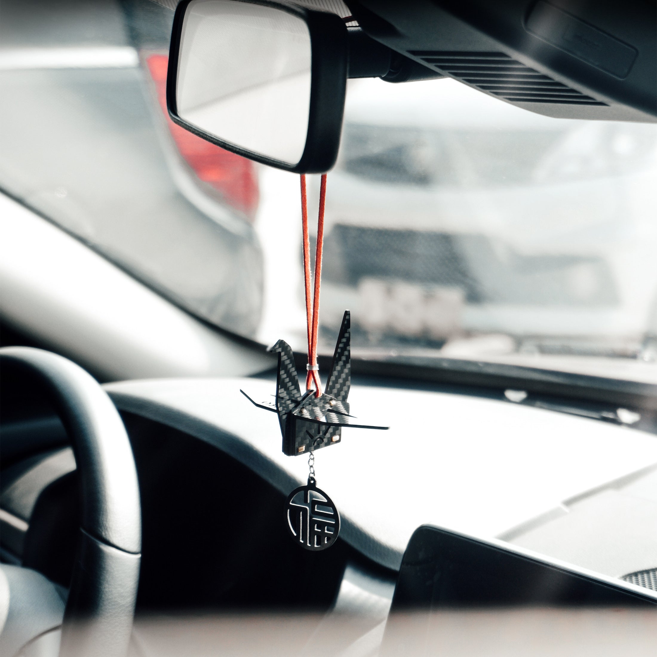 "Black carbon fiber-textured origami crane car charm hanging from a red cord on a vehicle’s rearview mirror; features a dangling circular pendant engraved with the Chinese 'good fortune' character, set inside a car (steering wheel and blurred exterior vehicle visible in background)"