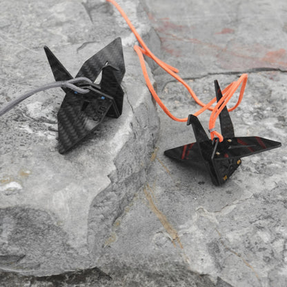 "Two black carbon fiber-textured origami crane accessories: one paired with a gray cord (featuring small blue accents) and the other with an orange cord (adorned with gold accents), placed on a textured gray rock surface"