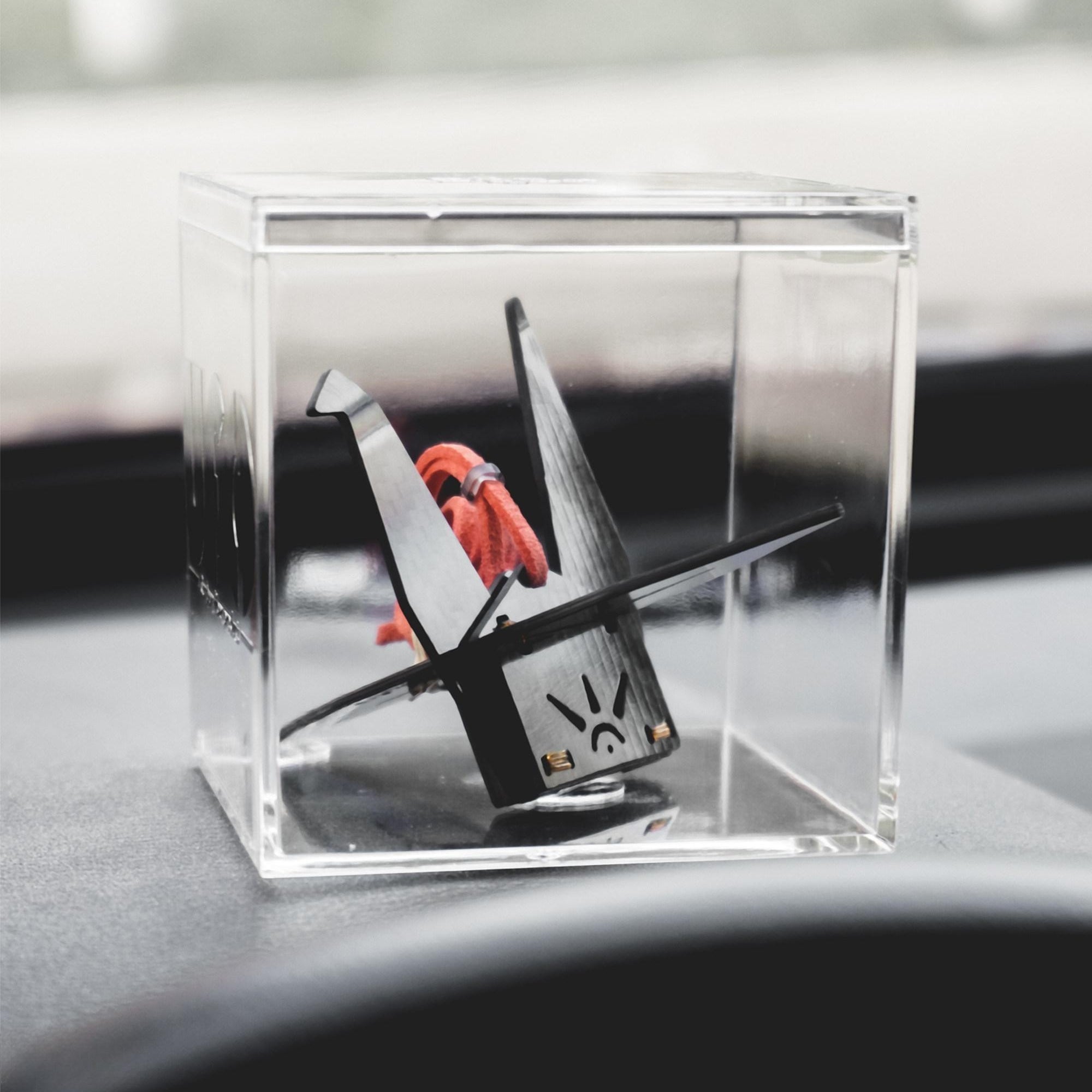 "Black carbon fiber-textured origami crane accessory (with an orange cord, sun-like design, and gold accents on its base) housed in a clear acrylic cube case, displayed on a car’s dashboard (softly blurred background in view)"