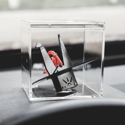 "Black carbon fiber-textured origami crane accessory (with an orange cord, sun-like design, and gold accents on its base) housed in a clear acrylic cube case, displayed on a car’s dashboard (softly blurred background in view)"