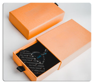 Two matte orange gift boxes (one open, one closed) placed on a light white surface: The open box features a black interior lining