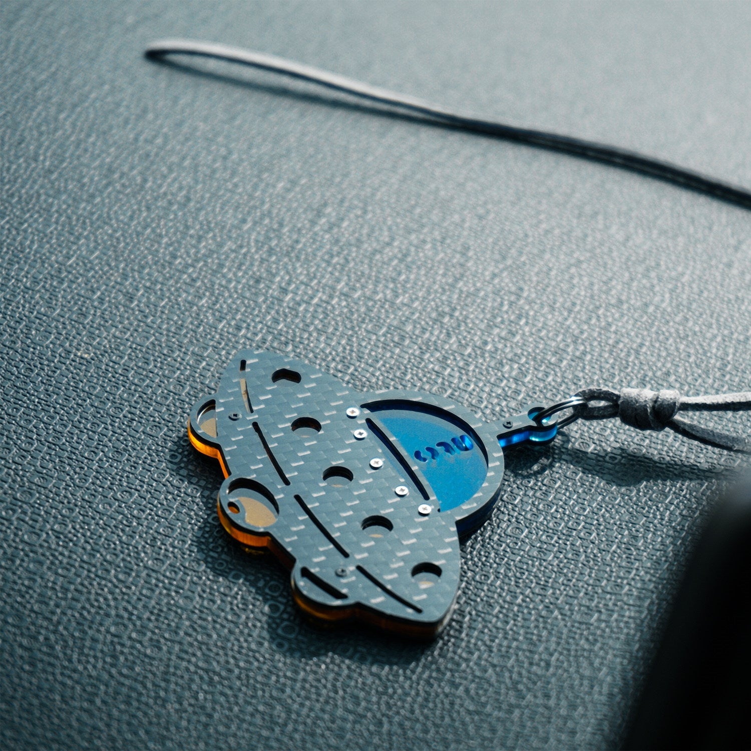 and small metallic rivets. The piece is attached to a gray leather cord (with metal hardware) and rests on a textured gray leather surface