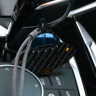 small metallic accents, and orange cutout details. Attached to a gray leather cord, the charm hangs near a car's gear shifter